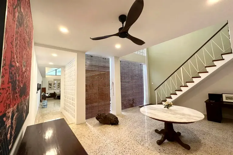 The double-height entry hall: terrazzo floor, timber chick blinds, a round marble-top table, the stair rising along an olive-green wall.